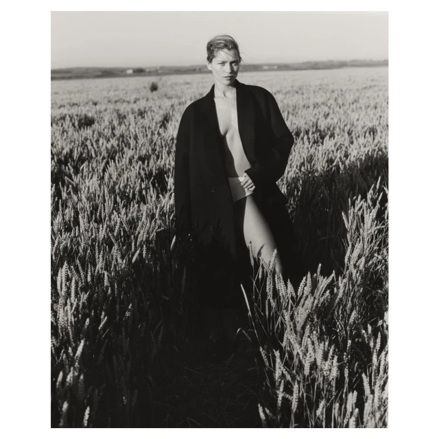 Black and white portrait of a woman in a coat standing in a wheat field, monochromatic and textural, with a rural outdoor setting.