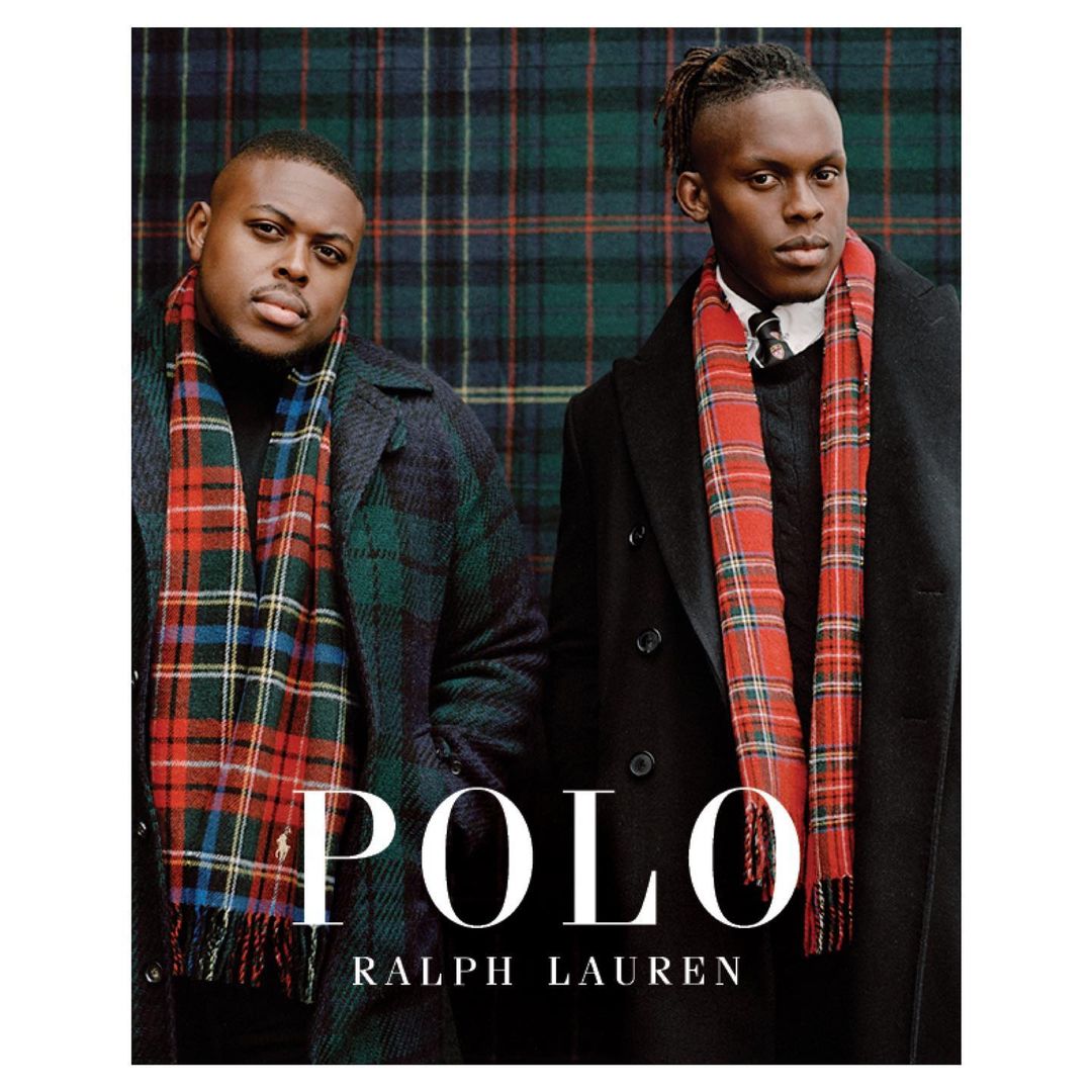 Two adult males stand indoors against a tartan backdrop, wearing dark overcoats and scarves in a fashion editorial by Polo Ralph Lauren.