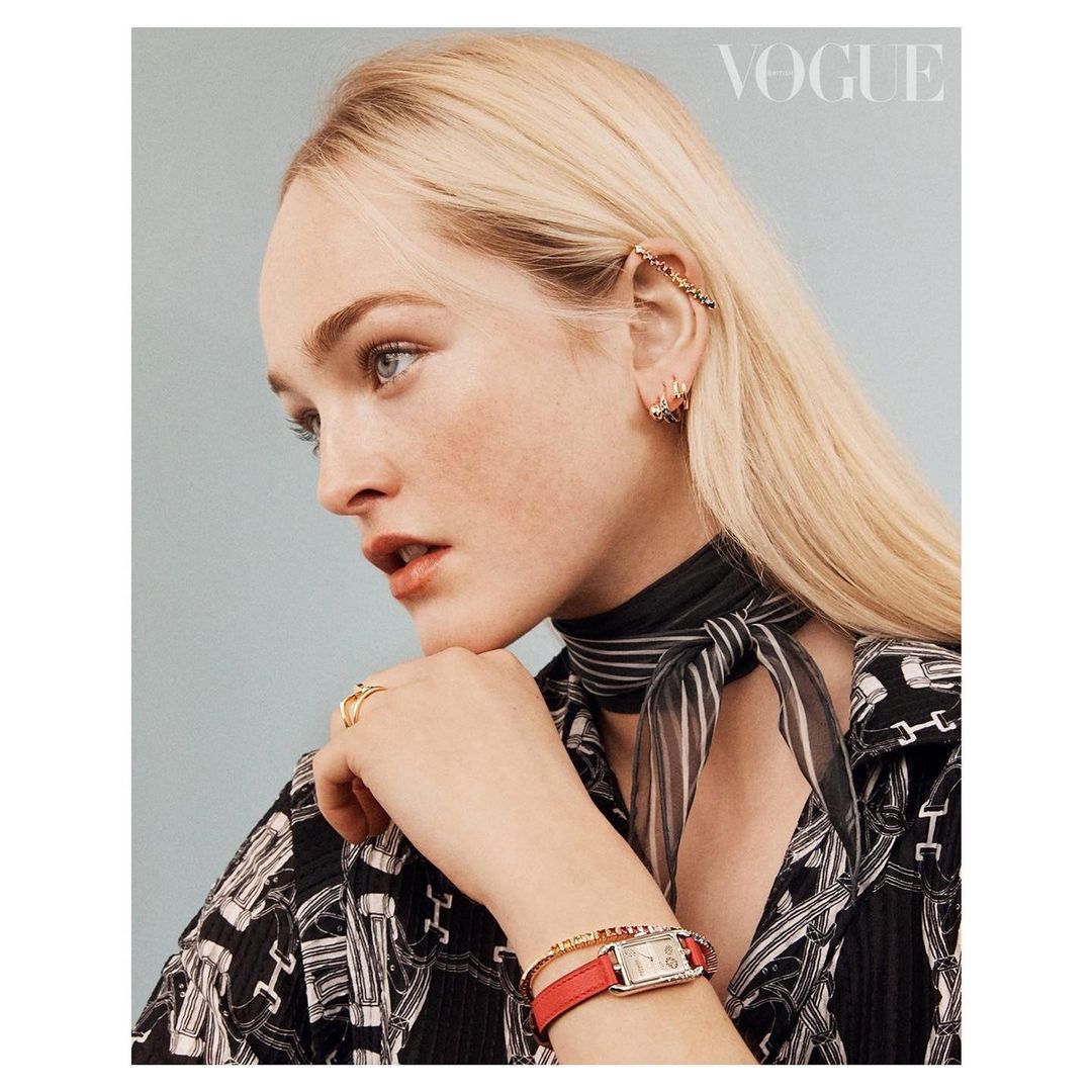 Close-up fashion shot of a blonde woman wearing jewelry, a red watch, and a patterned blouse in front of a light blue background.