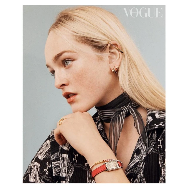 Close-up fashion shot of a blonde woman wearing jewelry, a red watch, and a patterned blouse in front of a light blue background.