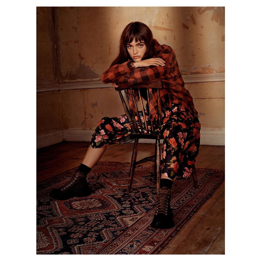 A young woman with bangs and a serious expression sits on a chair in a red and black plaid shirt and floral pants in a room.