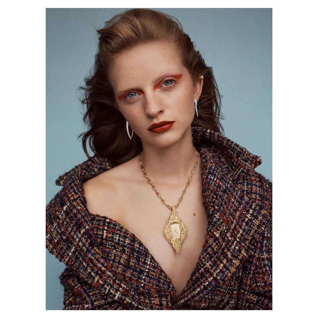Woman in a multicolor tweed jacket and statement necklace against a light blue backdrop, showcasing fashion, makeup, and jewelry details.
