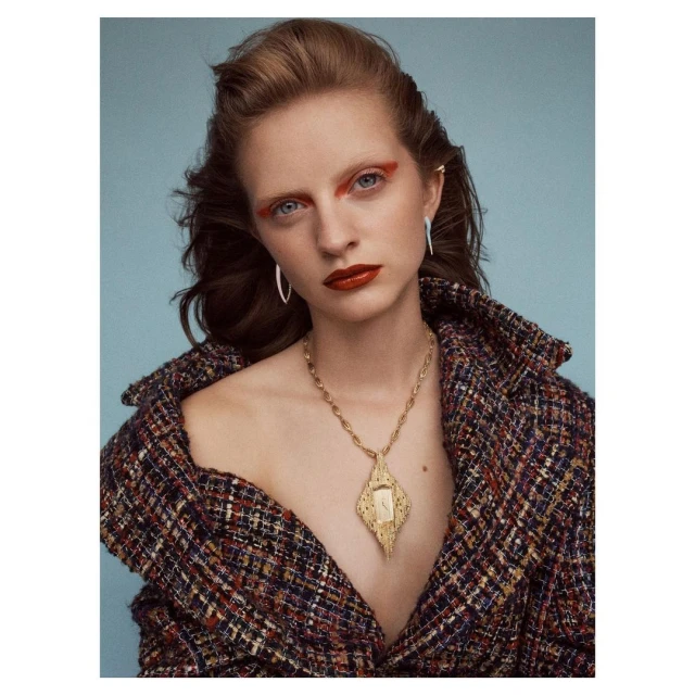 Woman in a multicolor tweed jacket and statement necklace against a light blue backdrop, showcasing fashion, makeup, and jewelry details.