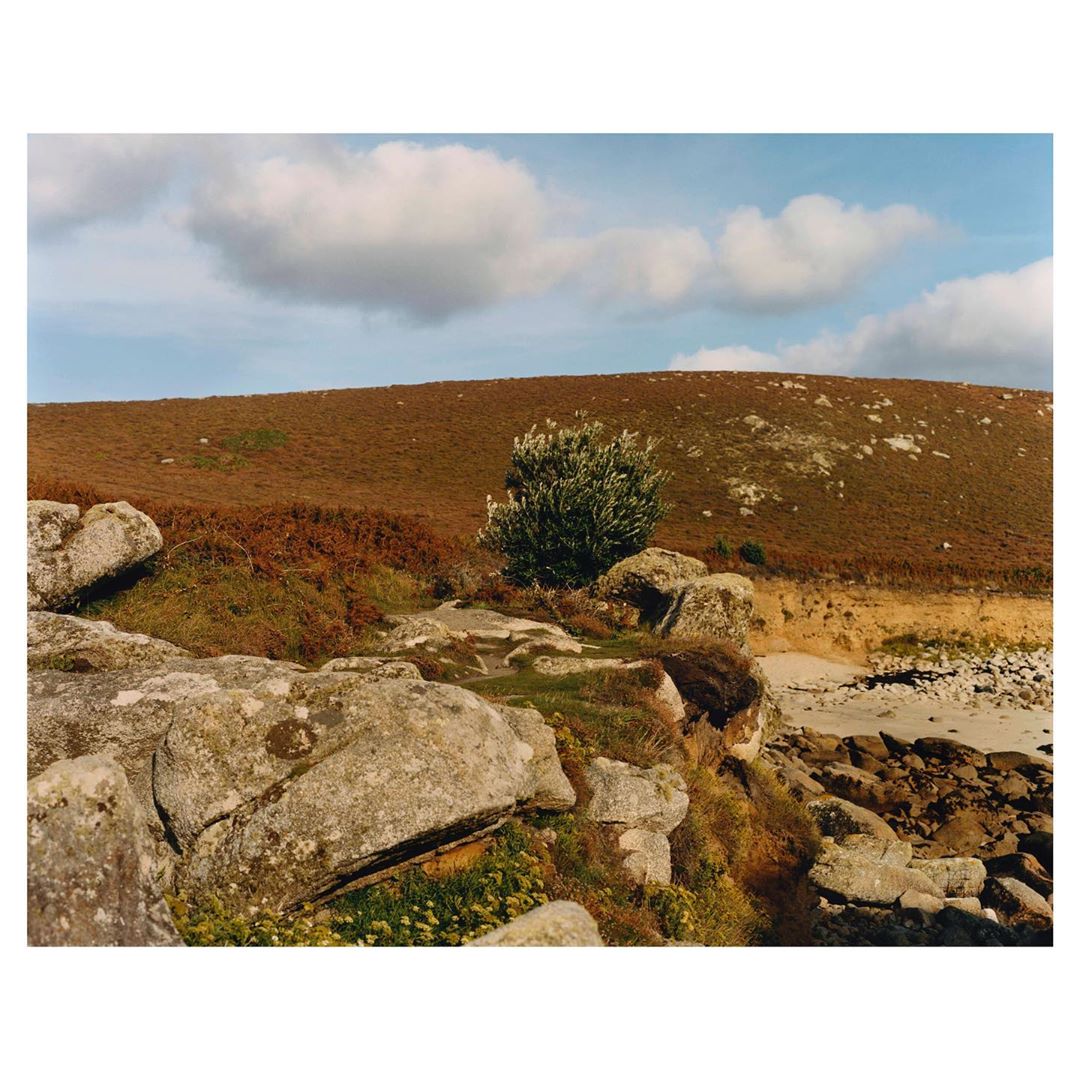 Scenic landscape with rocks and shrubbery in the foreground, a hill covered with brush in the background, and a partly cloudy blue sky.