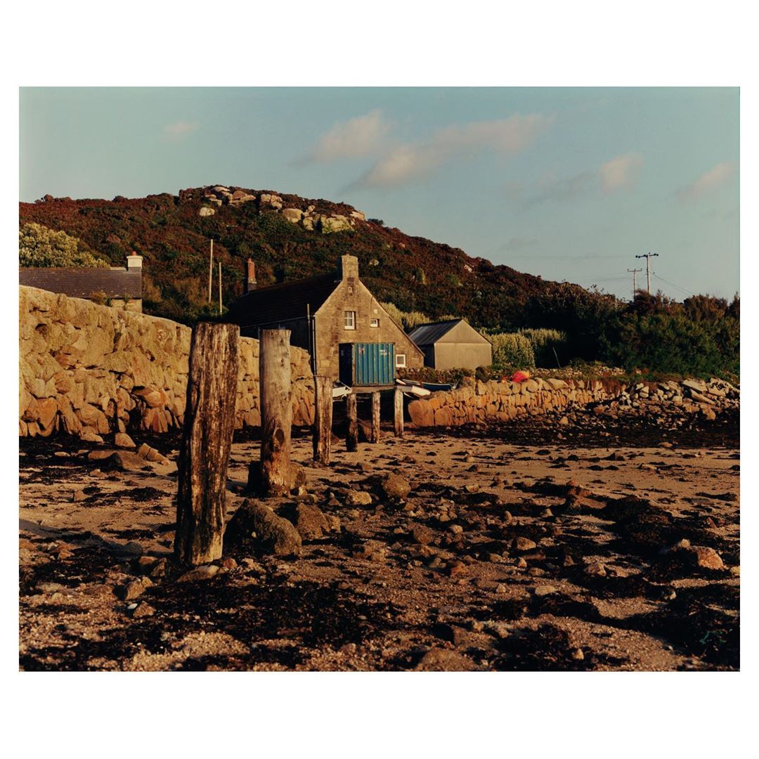 Exterior of a stone cottage on a rocky shoreline with wooden posts, featuring a hillside and pale blue sky, capturing a coastal landscape.