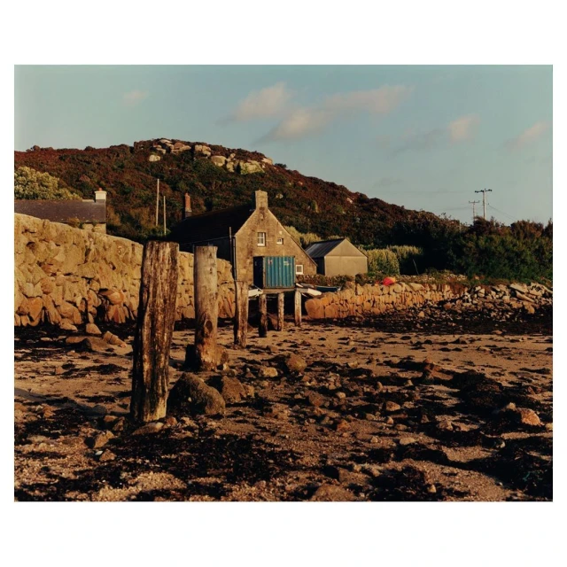 Exterior of a stone cottage on a rocky shoreline with wooden posts, featuring a hillside and pale blue sky, capturing a coastal landscape.