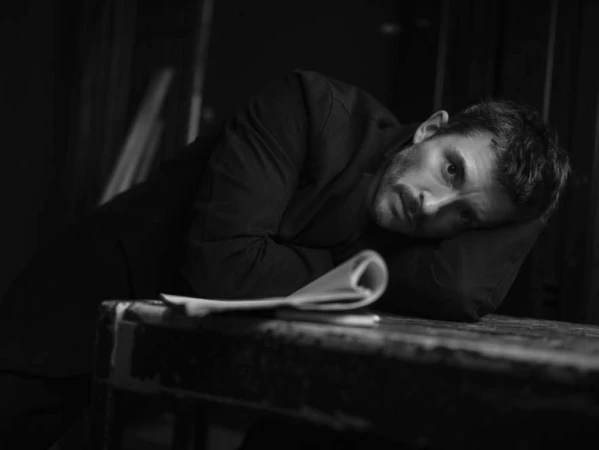 Monochrome portrait of a man resting his head on a wooden table beside a rolled document