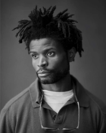 Black-and-white portrait of a man with dreadlocks wearing a polo shirt and glasses hung around his neck, photographed by Ben Weller.