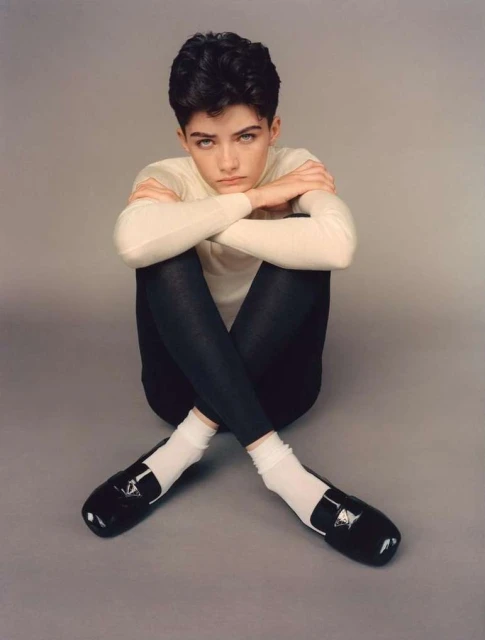 A young adult woman with short dark hair and neutral expression sits indoors wearing a cream sweater, dark leggings, white socks, and loafers.