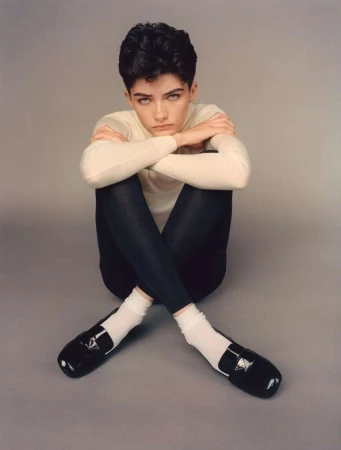 A young adult woman with short dark hair and neutral expression sits indoors wearing a cream sweater, dark leggings, white socks, and loafers.