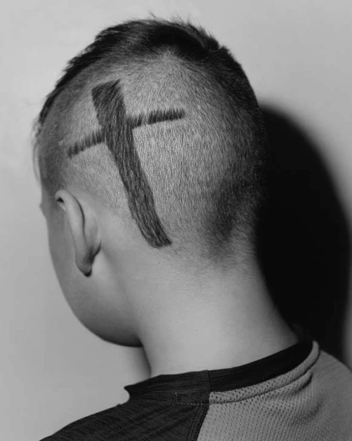 Close-up of the back of a child's head with a shaved haircut featuring a large cross design, set against a plain background.