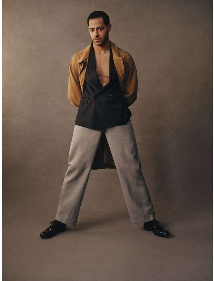 Man in a tan coat, black vest, and gray pants standing with legs spread in a neutral studio setting. Fashion and studio photography.