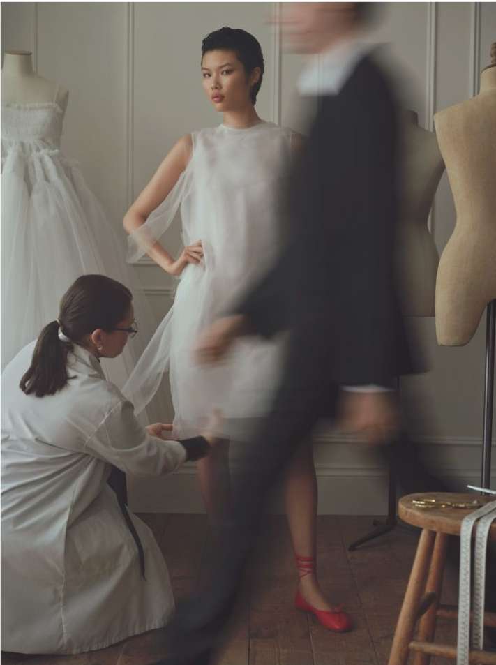 An Asian fashion model wears a semi-transparent white dress while a seamstress measures the hem in a light-filled photo studio.