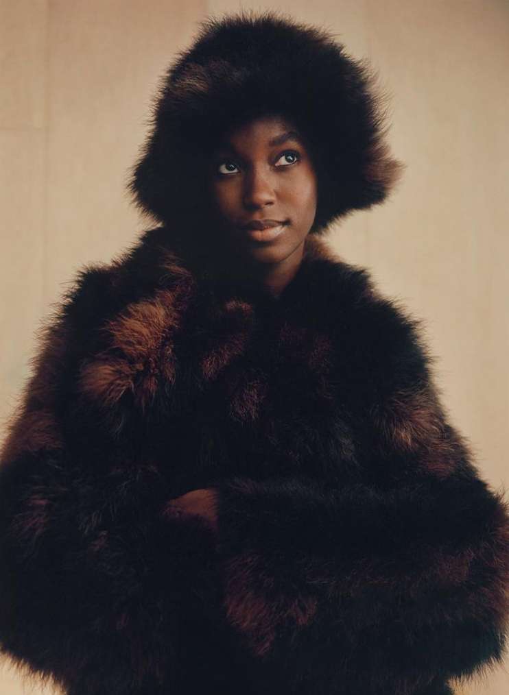 Portrait of a Black woman wearing a dark furry hat and matching textured coat against a warm neutral background.