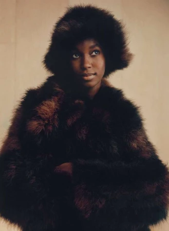 Portrait of a Black woman wearing a dark furry hat and matching textured coat against a warm neutral background.