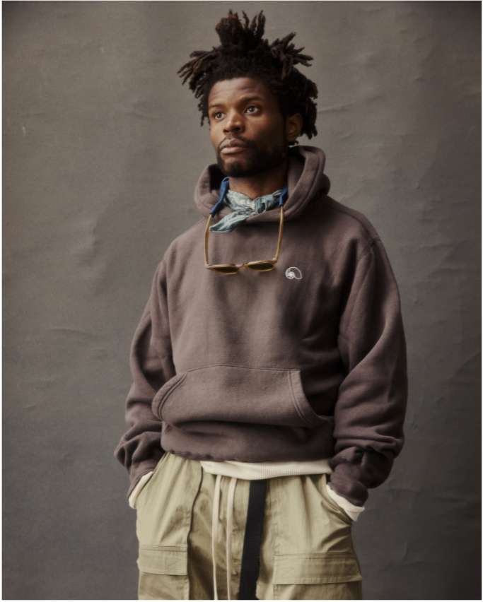 Portrait of a Black man wearing a brown hoodie and khaki pants against a gray backdrop, styled with sunglasses and a patterned scarf.