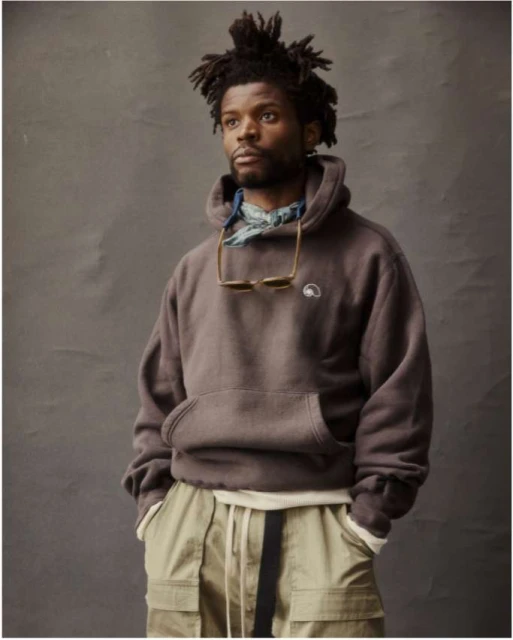 Portrait of a Black man wearing a brown hoodie and khaki pants against a gray backdrop, styled with sunglasses and a patterned scarf.