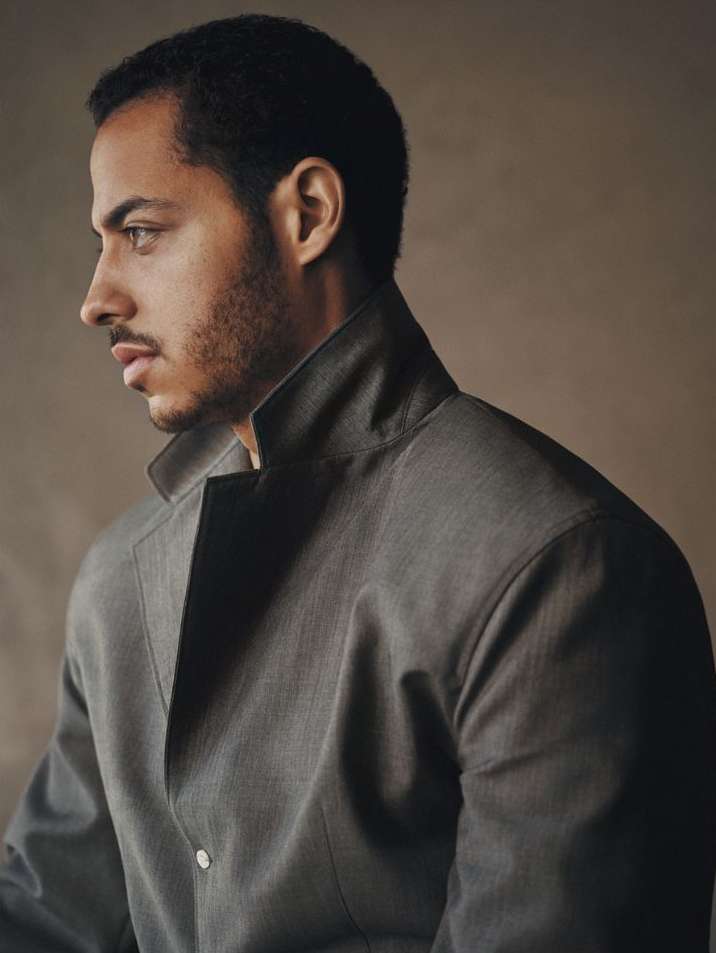 Portrait of a dark-haired man in a gray jacket looking to the left against a neutral background in a studio setting.