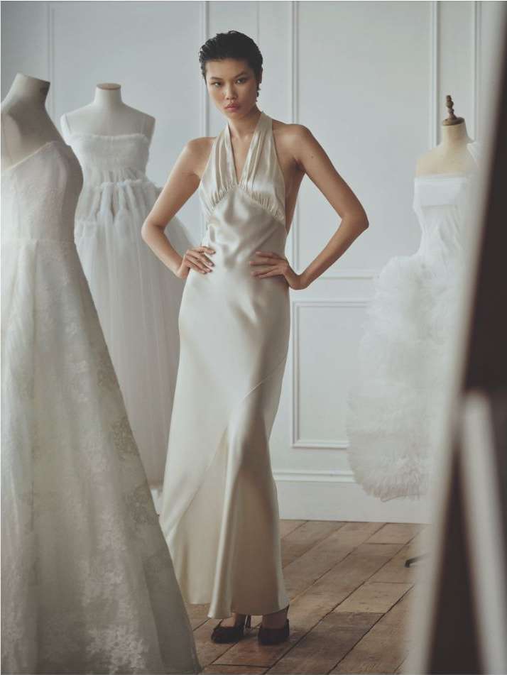 Woman in halter dress standing in an indoor fashion studio surrounded by dress forms wearing white gowns in a fashion editorial setting.