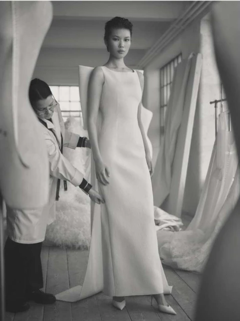 A stylish, elegant black and white fashion shot featuring a woman in a long white dress, being fitted by a tailor in a workshop.