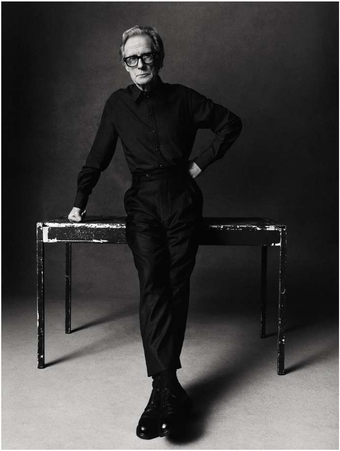 Black and white portrait of an adult male with glasses, posing near a table in a studio setting, wearing dark clothing with dress shoes.