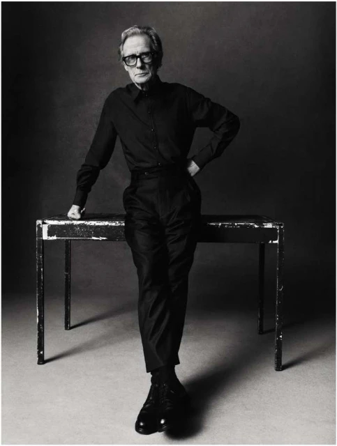 Black and white portrait of an adult male with glasses, posing near a table in a studio setting, wearing dark clothing with dress shoes.