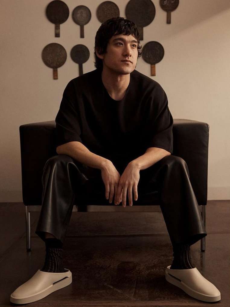 Portrait of a stylish Asian man in casual clothing sitting in a black chair with decoratively arranged, round, antique mirrors on the wall.
