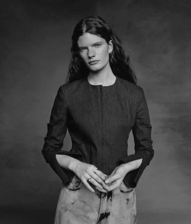Monochrome portrait of a woman wearing a structured dark jacket and light patterned jeans against a textured backdrop
