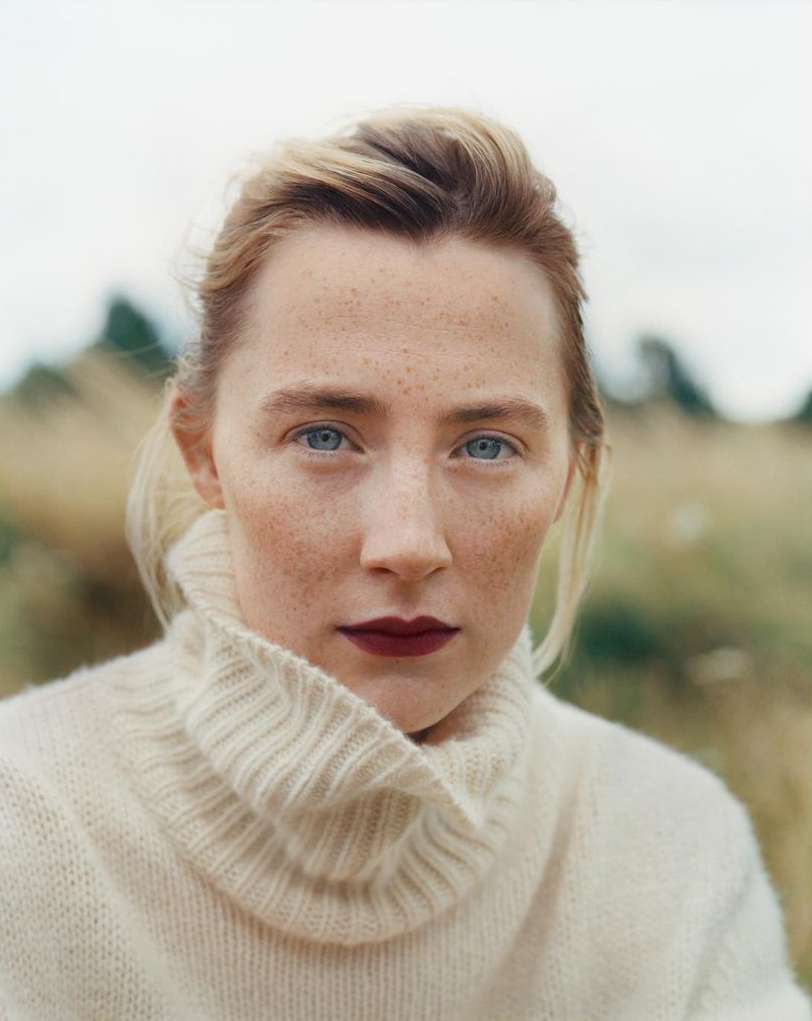 Close-up portrait of a freckled woman with blue eyes and a neutral expression wearing a cream-colored turtleneck sweater in a grassy field.