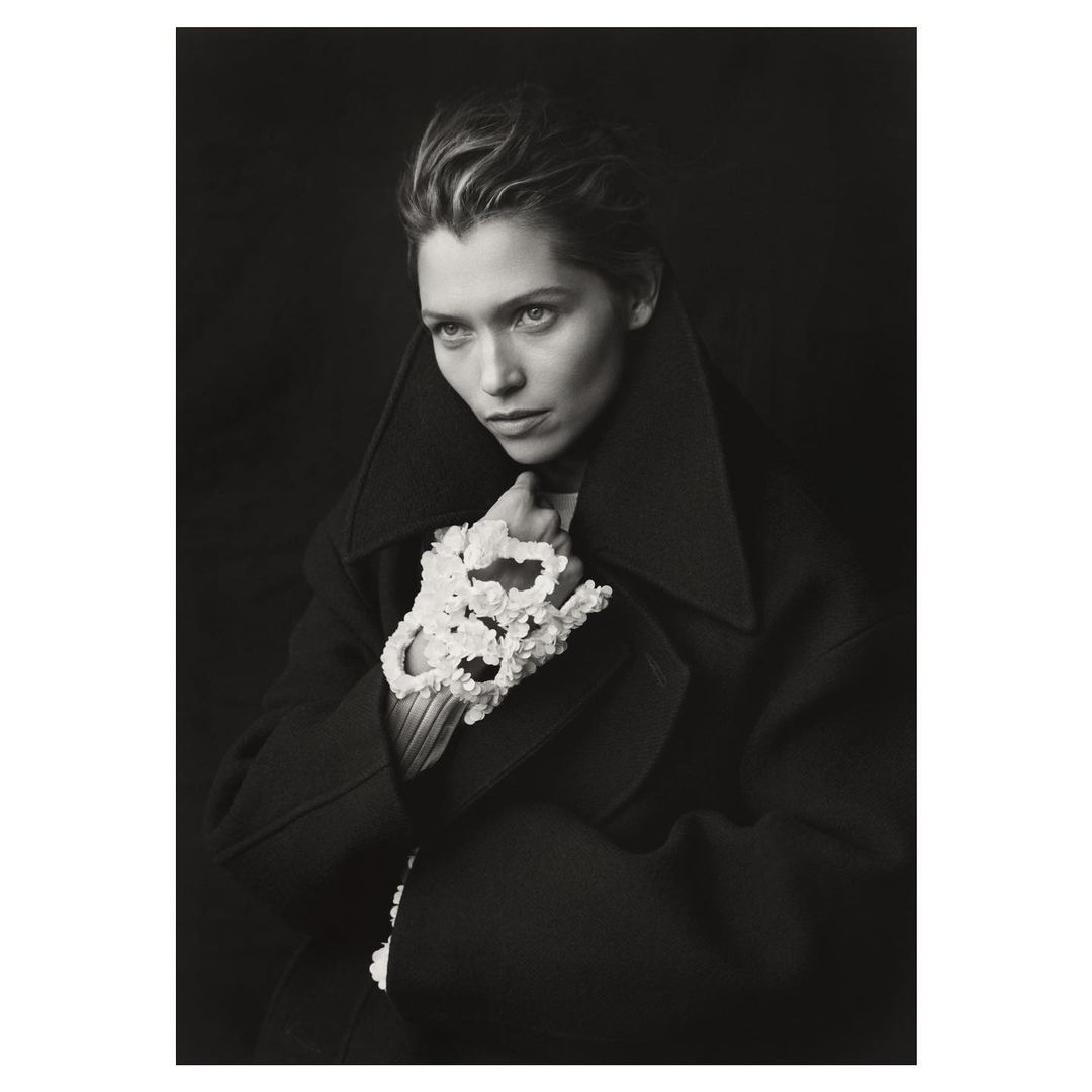 Portrait of a woman in a dark coat with sculptural floral gloves, gazing slightly away, set against a dark studio backdrop.