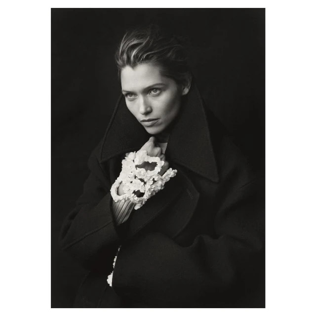 Portrait of a woman in a dark coat with sculptural floral gloves, gazing slightly away, set against a dark studio backdrop.