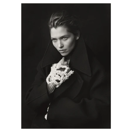 Portrait of a woman in a dark coat with sculptural floral gloves, gazing slightly away, set against a dark studio backdrop.