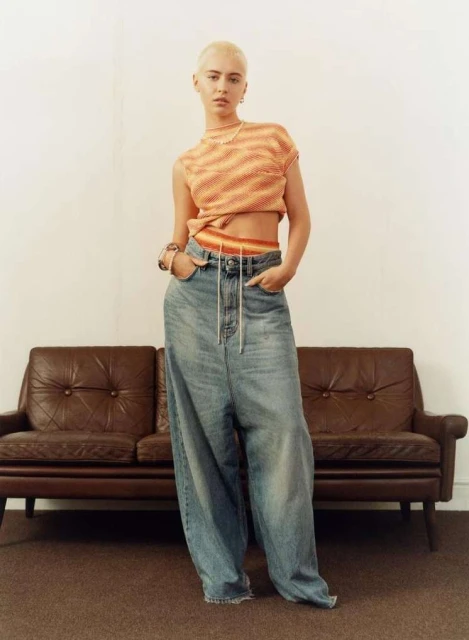 A woman with short hair poses in a striped, cropped sweater and oversized denim pants against a neutral backdrop with a brown couch.