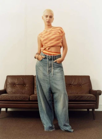 A woman with short hair poses in a striped, cropped sweater and oversized denim pants against a neutral backdrop with a brown couch.
