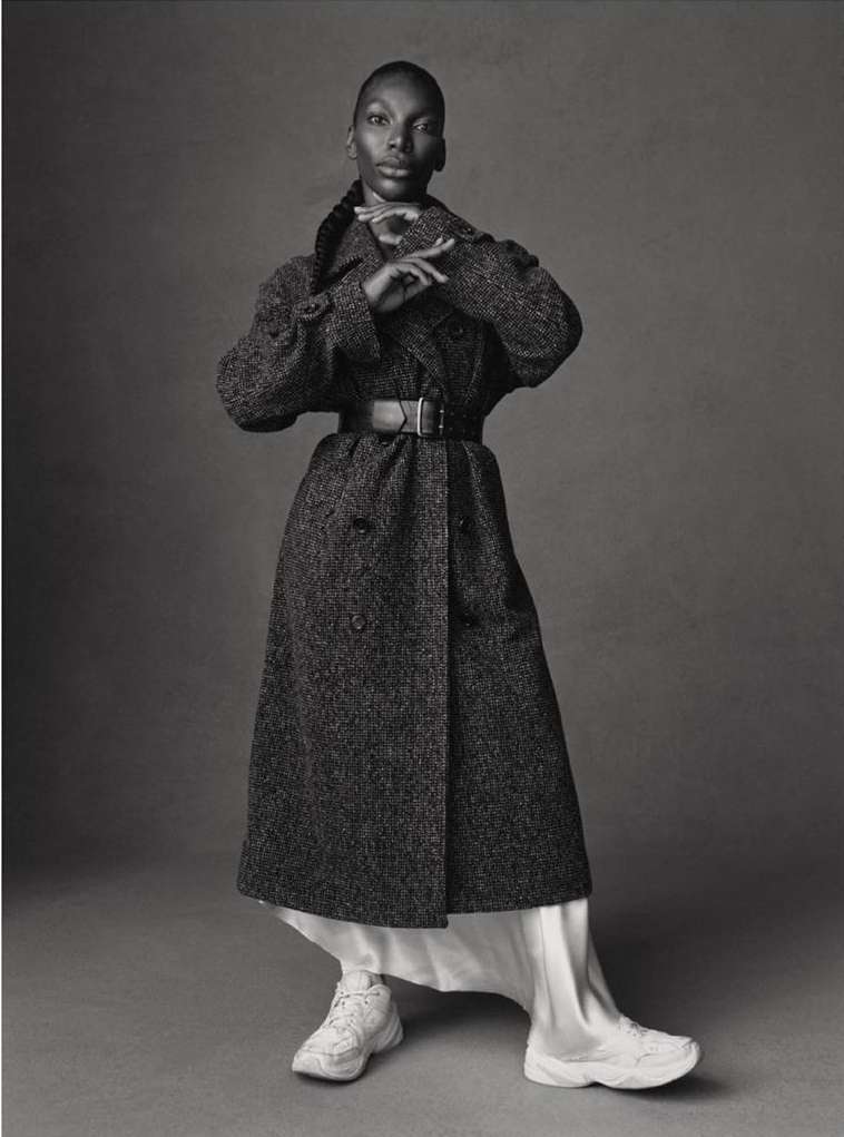 Full-length studio shot of a Black female model in a long, tweed coat, white dress, and sneakers against a grey backdrop.