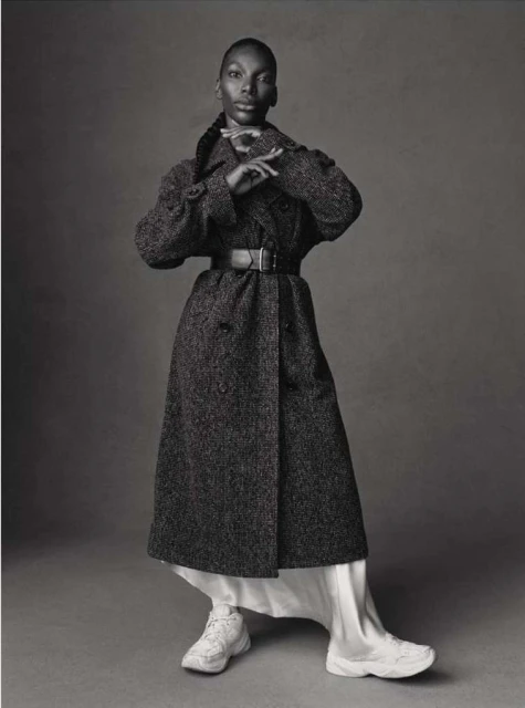 Full-length studio shot of a Black female model in a long, tweed coat, white dress, and sneakers against a grey backdrop.