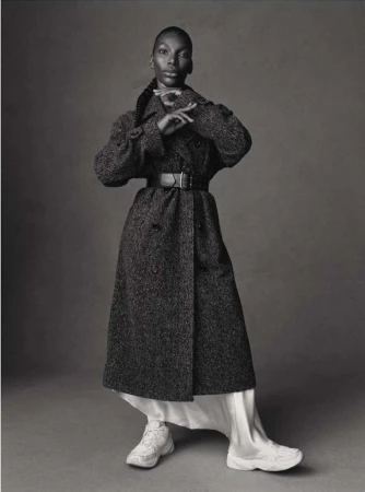 Full-length studio shot of a Black female model in a long, tweed coat, white dress, and sneakers against a grey backdrop.