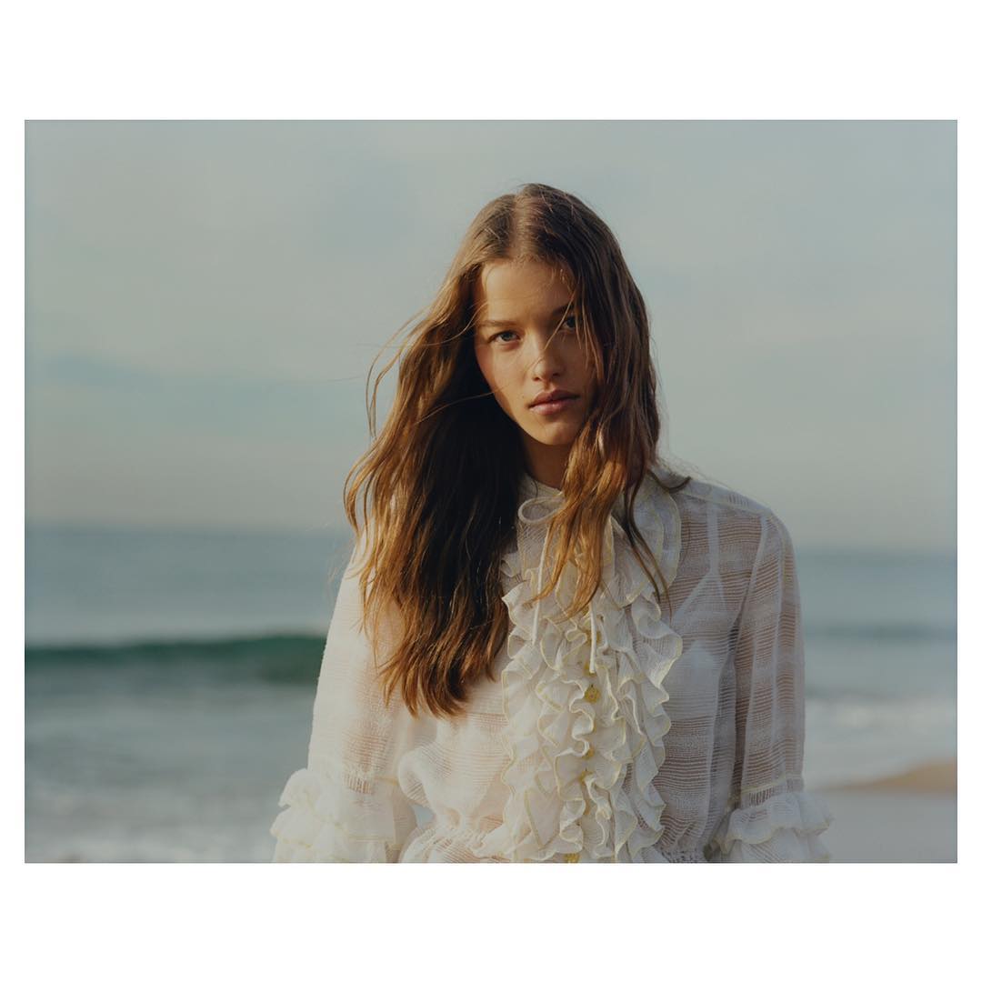 A portrait of a young woman with long brown hair stands on a beach in front of the ocean wearing a white frilly blouse.