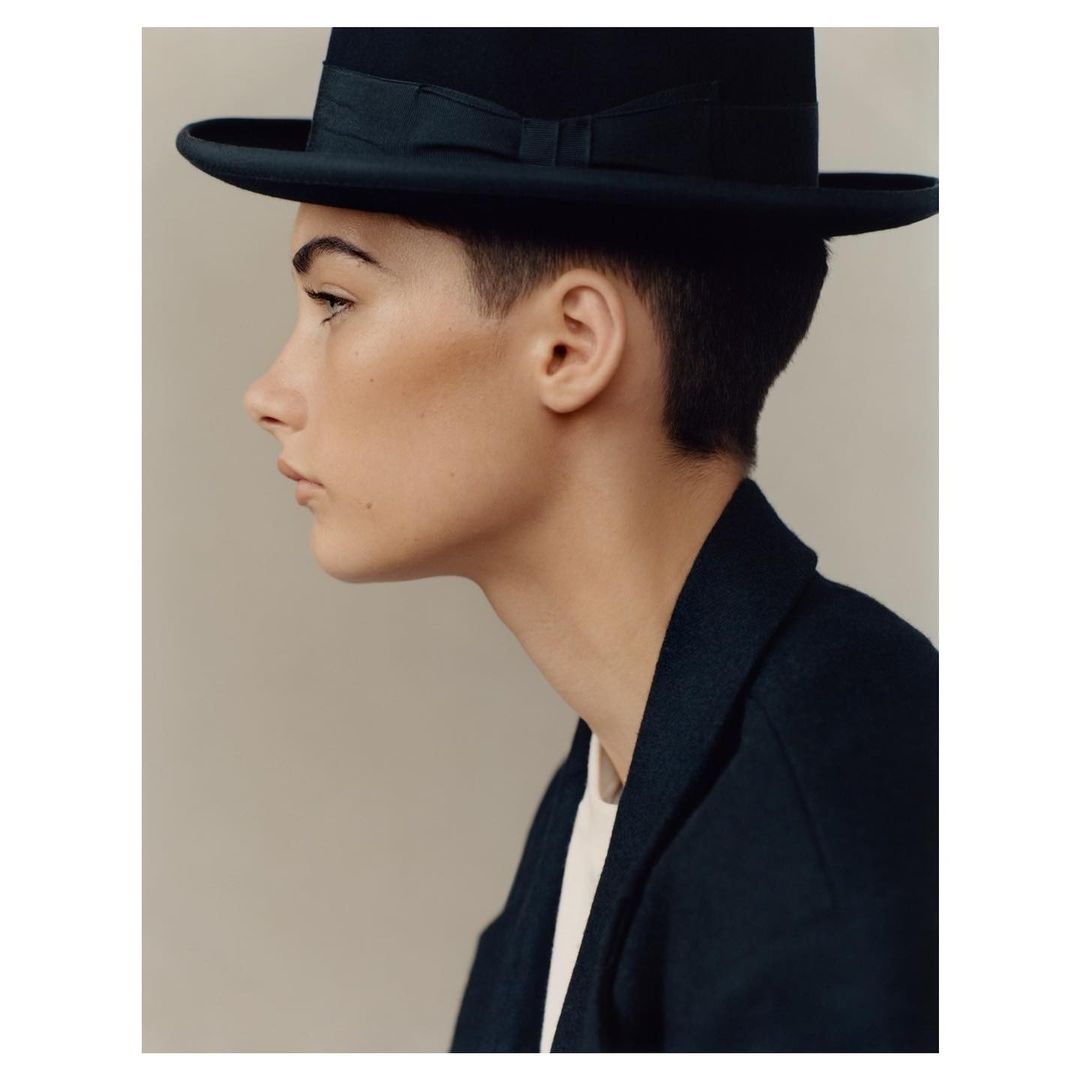 Profile portrait of an adult female wearing a black fedora hat and a dark blazer in a studio setting with neutral background.