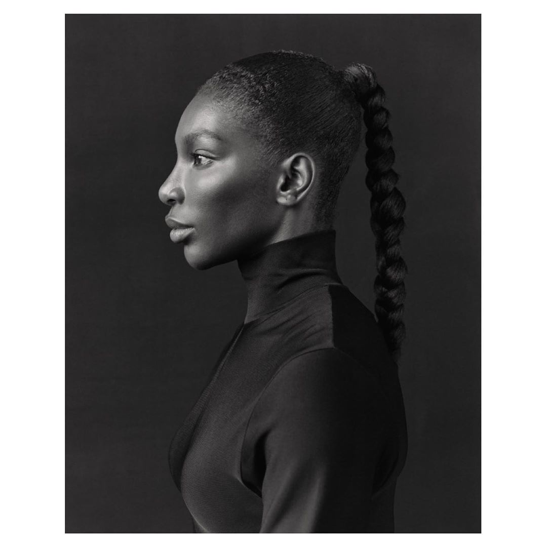 Monochrome portrait of a Black woman in profile with braided ponytail, wearing a turtleneck against a dark gray background.