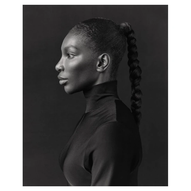 Monochrome portrait of a Black woman in profile with braided ponytail, wearing a turtleneck against a dark gray background.