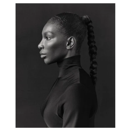 Monochrome portrait of a Black woman in profile with braided ponytail, wearing a turtleneck against a dark gray background.