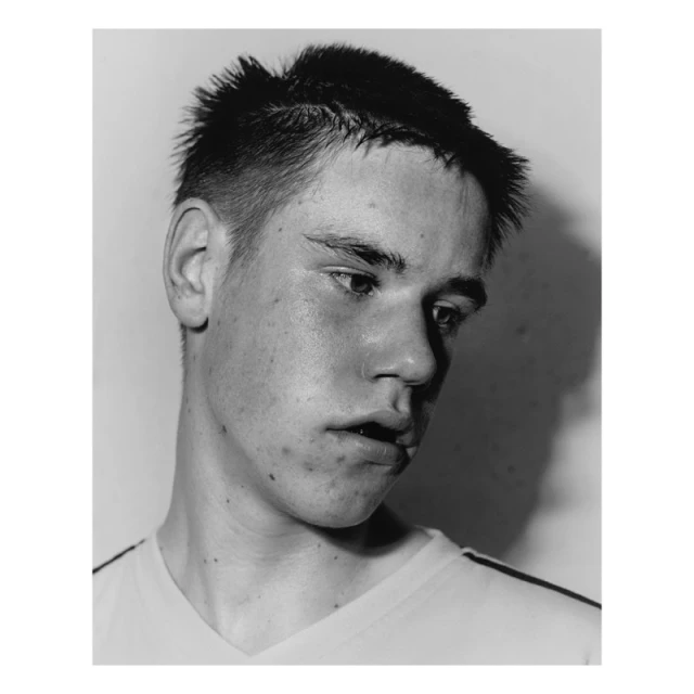 Monochrome portrait of a young adult male with short spiky hair, looking down, wearing a v-neck shirt against a neutral background.