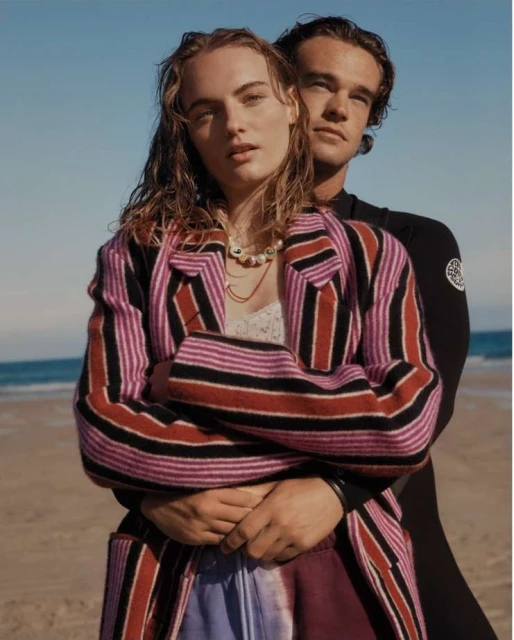A young couple poses on a beach. The woman wears a striped jacket and the man wears a wetsuit; the ocean and sky are visible in the background.