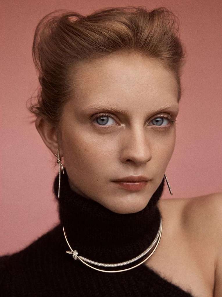 Close-up portrait of a woman wearing a black turtleneck and silver jewelry against a muted pink background.