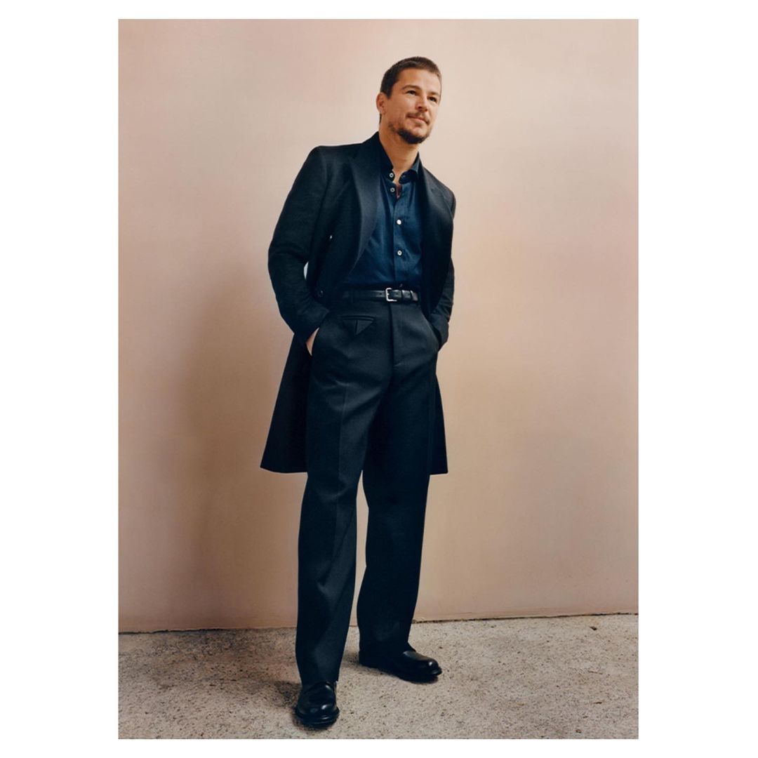 Full-length shot of adult man in dark blue shirt, black trousers, black shoes, and a dark coat standing indoors against a neutral wall.
