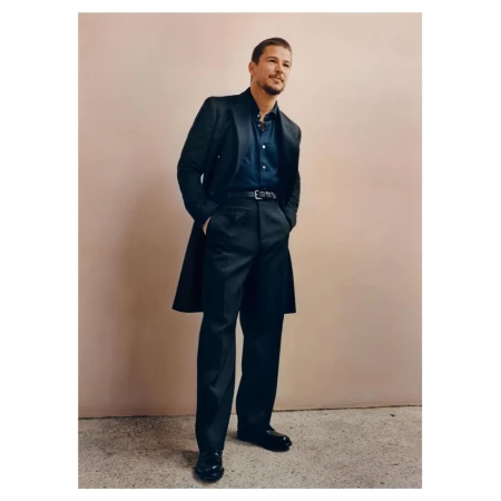 Full-length shot of adult man in dark blue shirt, black trousers, black shoes, and a dark coat standing indoors against a neutral wall.