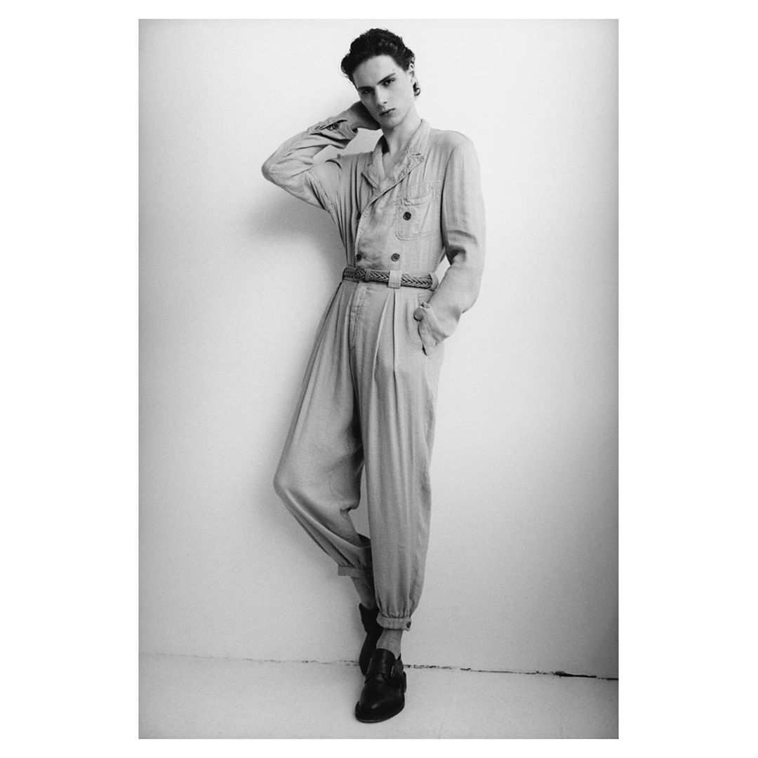 Black and white fashion portrait of a male model in a casual, buttoned jumpsuit and loafers posing against a white wall.