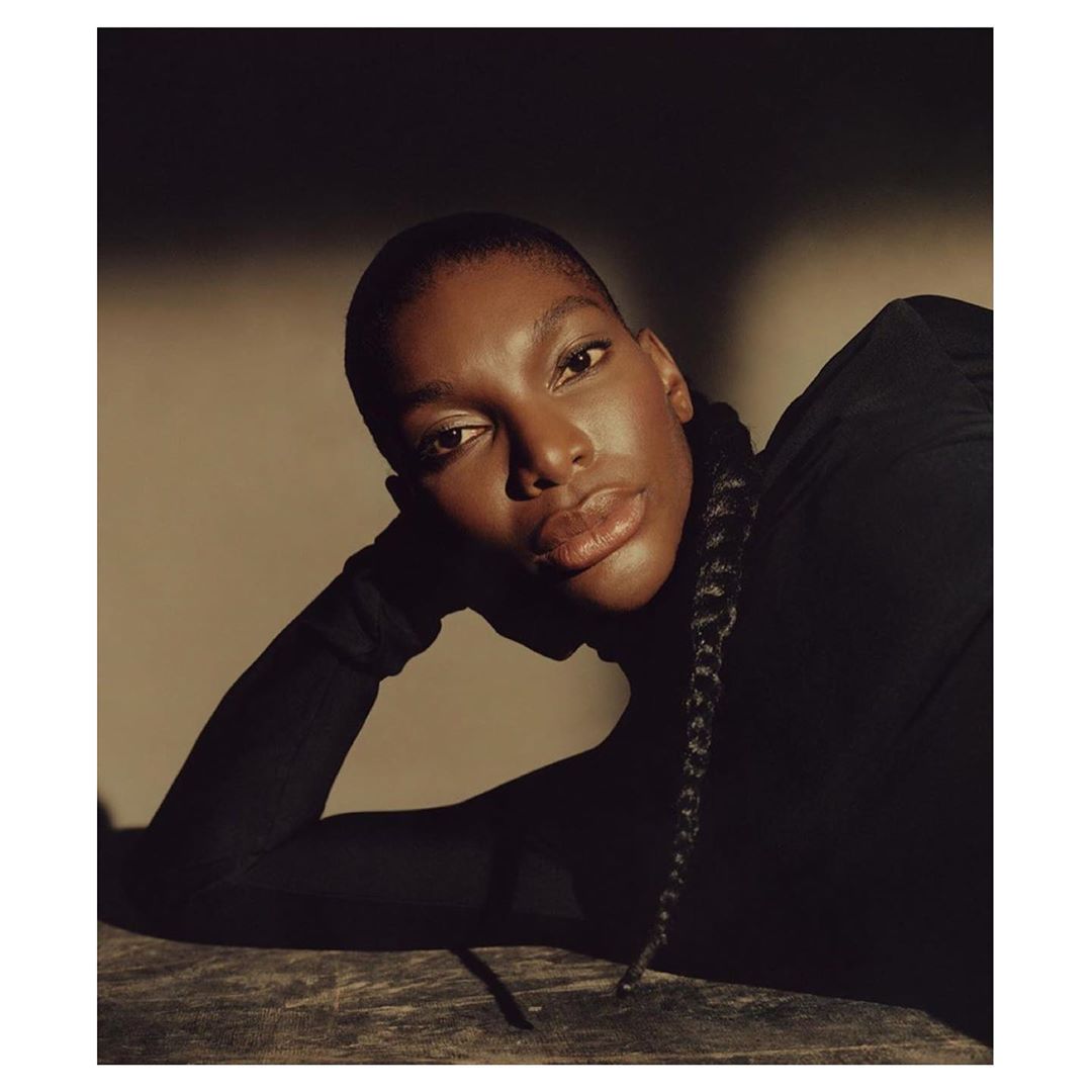 Close-up portrait of a dark-skinned woman with short hair wearing a black turtleneck and posing with a neutral facial expression.