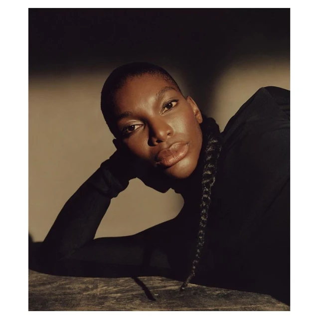 Close-up portrait of a dark-skinned woman with short hair wearing a black turtleneck and posing with a neutral facial expression.